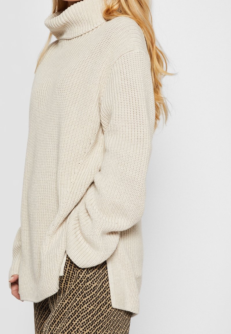 Femme portant un pull à col roulé en grosse maille couleur crème avec des manches longues et un pantalon imprimé animal, debout devant un fond uni.