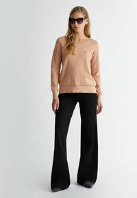 Maglione a maglia pesco con polsini e orlo a costine; jeans neri a zampa d'elefante; occhiali da sole scuri completano l'outfit. L'abbigliamento presenta un dettaglio con tasca.