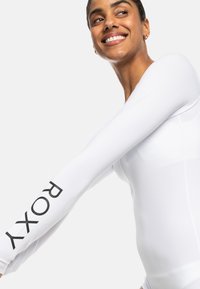 White long-sleeve rash guard with a fitted design, made of smooth material, featuring a large black "ROXY" logo on the left sleeve.