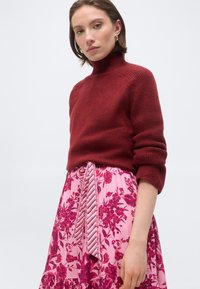 Γυναίκα που φοράει πουλόβερ μπορντό με λαιμόκοψη ζιβάγκο και μανίκια που έχουν τυλιχτεί, καθώς και ροζ φλοράλ φούστα με ζώνη από υφασμάτινες ρίγες.