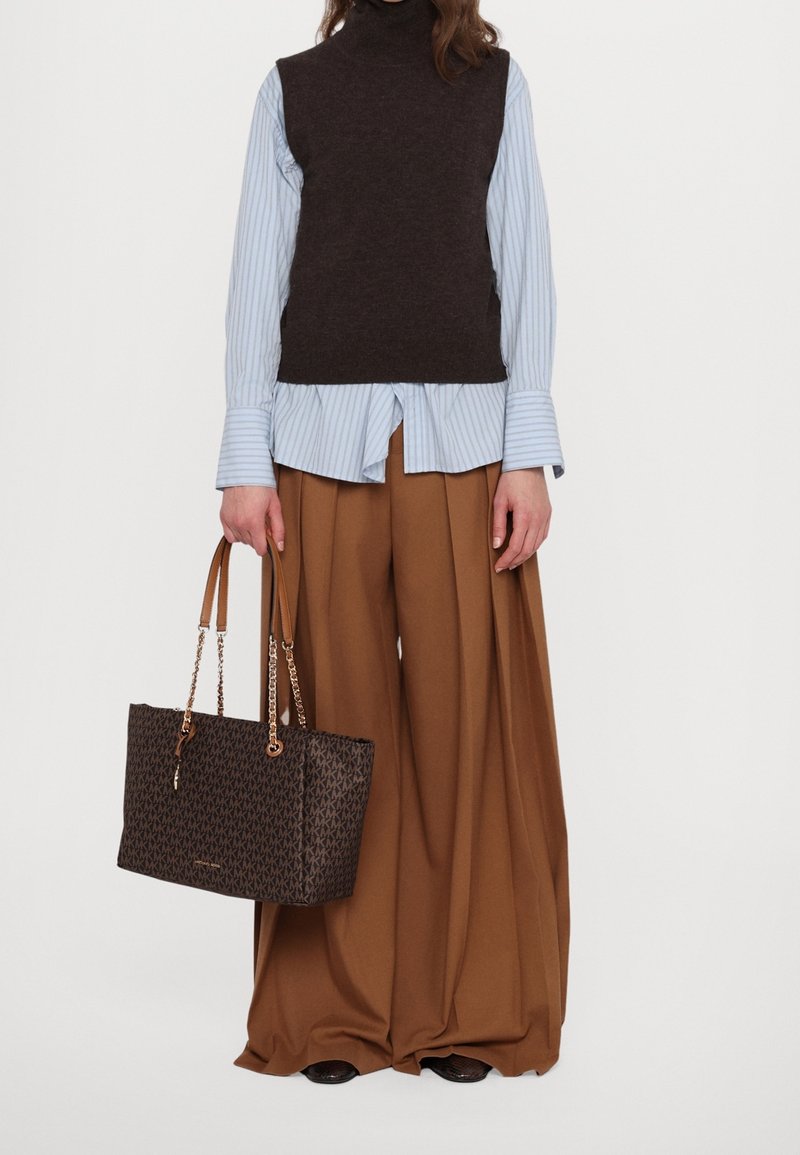 Personne portant un pull sans manches noir par-dessus une chemise rayée bleue avec un pantalon large marron, tenant un sac cabas marron foncé.