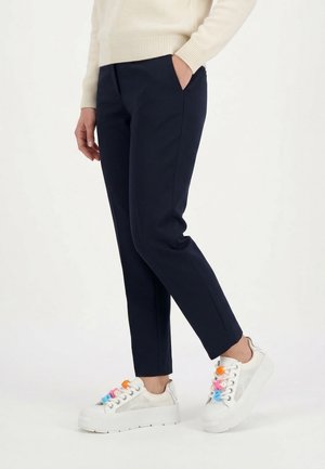 Person trägt marineblaue, schmal zulaufende Hose und weiße Plateausneaker mit mehrfarbigen Schnürsenkeln, steht vor einem einfarbigen Hintergrund.