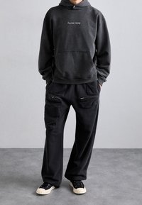 Sudadera negra con capucha, bolsillo frontal y logo, combinada con pantalones negros holgados con bolsillos con cremallera. Zapatillas negras con suelas blancas.