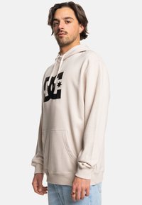Beige hoodie made of soft fabric; features a large black DC logo on the front, a kangaroo pocket, and ribbed cuffs and hem.