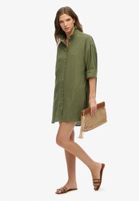 Vestido de camisa verde de lino con botones, mangas enrolladas, acompañado de un clutch tejido beige y sandalias deslizantes marrones con acentos circulares.
