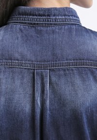Visión trasera de una persona que lleva una camisa de mezclilla azul mediana con costuras detalladas en el cuello y la yugo, con el cabello recogido detrás del cuello.