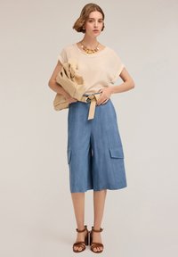 Blusa beige chiaro con maniche arrotolate, abbinata a culottes azzurri chiari con grandi tasche laterali e una cintura beige, completata da sandali marroni.
