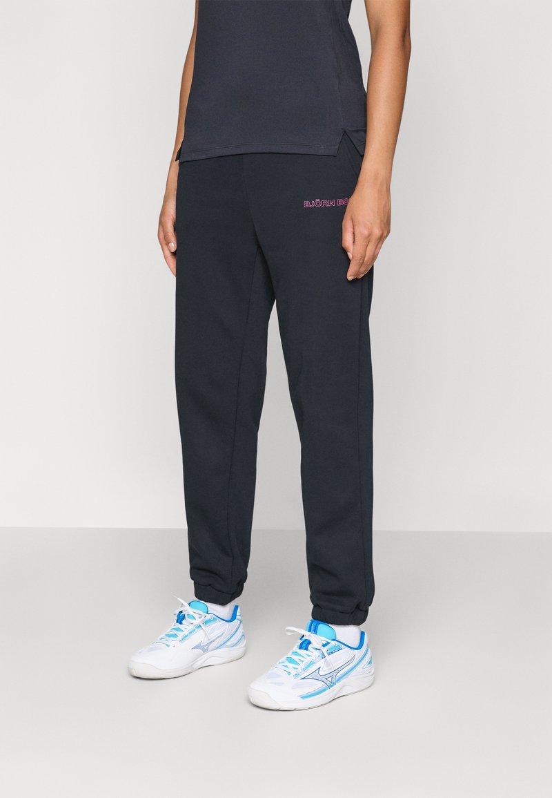 Tmavošedé bavlnené jogger nohavice s elastickými manžetami a malým ružovým logom na stehne, v kombinácii s bielymi teniskami s modrými akcentmi.