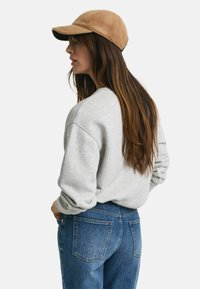 Grijze sweatshirt met verlaagde schouders, gecombineerd met blauwe denim jeans. Een bruin petje maakt de look af, met een geborduurd logo aan de voorkant.