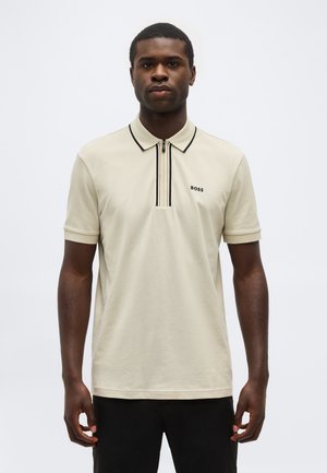 Mand iført beige kortærmet poloshirt med sort lynlås og kravekant, stående mod en ensartet lys baggrund.