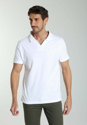 Homme aux cheveux courts et barbe taillée, portant un polo blanc et un pantalon olive, regardant légèrement vers sa gauche dans un environnement neutre.