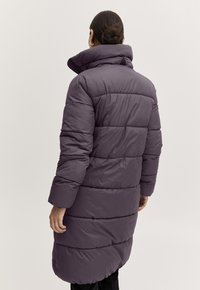 Kobieta z ciemnymi włosami upiętymi w kok, ubrana w długi, pikowany, ciemnofioletowy zimowy płaszcz z wysokim kołnierzem, stojąca plecami na jednolitym tle.