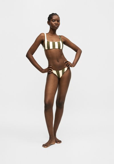 Γυναίκα στέκεται με τα χέρια στη μέση, φορώντας ένα μπικίνι με κάθετες πράσινες και λευκές ρίγες σε μονόχρωμο ανοιχτό φόντο.