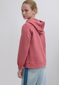 Pink cotton hoodie with a drawstring hood, ribbed cuffs, and hem. Paired with light blue jeans featuring a panel design.
