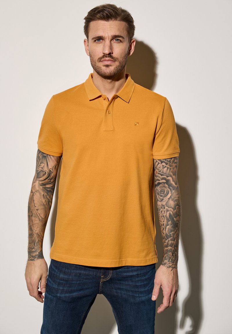 Musztardowa polo z miękkiego materiału, z klasycznym kołnierzykiem i trzema guzikami, zestawiona z ciemnoniebieskimi dżinsami.