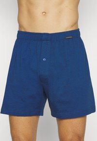 Boxers bleus pour hommes, en tissu tissé, avec une taille élastique, une fermeture à un bouton, et une texture subtile avec une petite étiquette logo sur la taille.