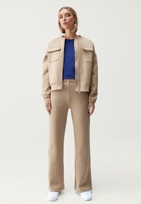 Beige bomberjacka med två framfickor, en marinblå topp och beiga vida byxor. Modellen bär vita skor; slät, texturerad tyg.