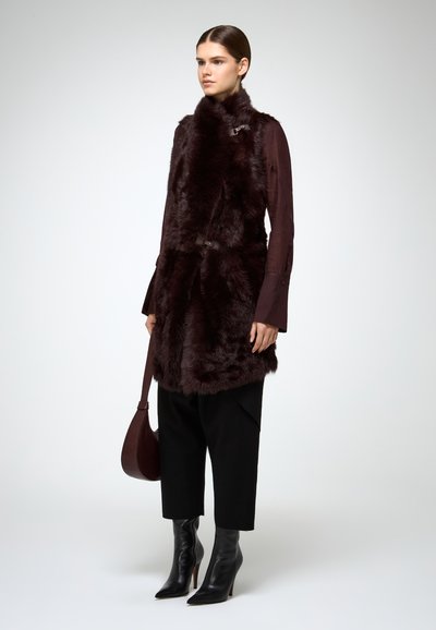 Donna in piedi con un gilet di pelliccia marrone scuro, camicia a maniche lunghe, pantaloni neri a cropped, stivali neri con tacco, mentre tiene una borsa marrone abbinata contro uno sfondo bianco.
