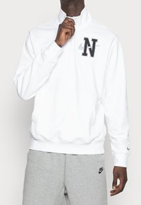 Sudadera blanca con media cremallera, hecha de una tela suave, que cuenta con un logo negro de "N" y un pequeño tick de Nike. Combinada con pantalones de chándal grises.