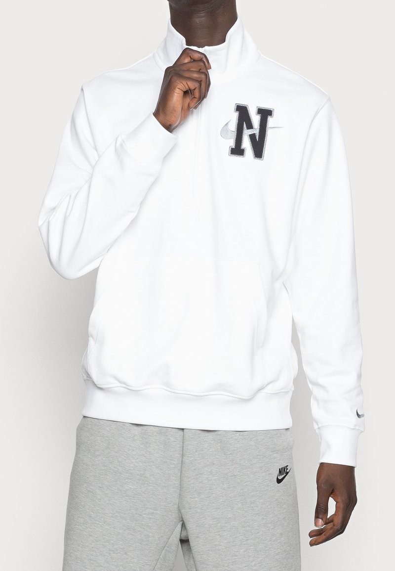 Sudadera blanca con media cremallera, hecha de una tela suave, que cuenta con un logo negro de "N" y un pequeño tick de Nike. Combinada con pantalones de chándal grises.