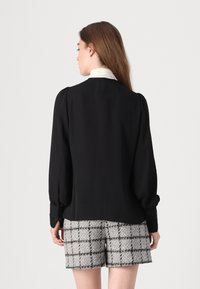 Svart bluse med puffede ærmer og høj krave, parret med beige ternede shorts. Blusen har blonde detaljer ved ærmerne. Glat stof.