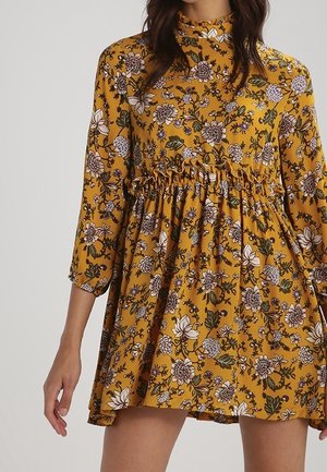 Kvinde iført en sennepsgul kjole med hvidt og lilla blomsterprint og indsnøret talje, med trekvartlange ærmer.