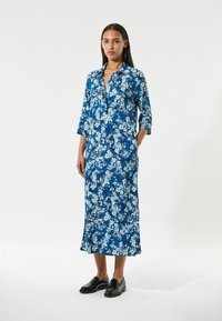 Blaues, langes Kleid mit floralem Muster und Dreiviertelärmel, Kragen und seitlichen Schlitzen, kombiniert mit schwarzen Slippern, getragen von einem Model vor einem schlichten Hintergrund.