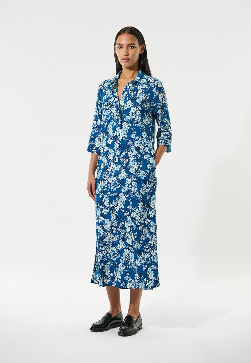 Blaues, langes Kleid mit floralem Muster und Dreiviertelärmel, Kragen und seitlichen Schlitzen, kombiniert mit schwarzen Slippern, getragen von einem Model vor einem schlichten Hintergrund.