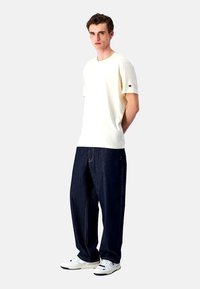 Weißes Kurzarm-T-Shirt mit einem Logo auf dem linken Ärmel, kombiniert mit lockeren, dunkelblauen Jeans und weißen Sneakers.
