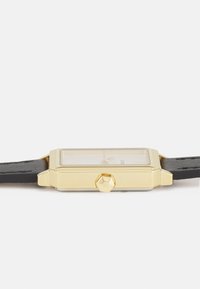 Montre rectangulaire en or avec un cadran transparent, bracelet en cuir noir et couronne texturée. Aiguilles des heures et des minutes simples visibles.