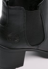 Bottine noire en cuir avec un panneau latéral élastique, un talon bloc, une languette de tirage et un logo circulaire embossé près du talon.