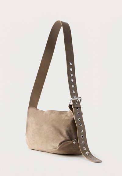 Μαλακή μπεζ τσάντα ώμου από σουέντ με φαρδιά ρυθμιζόμενη τιράντα που έχει πολλαπλές ασημένιες τρύπες και ασημένια αγκράφα, εμφανίζεται σε απλό φόντο.