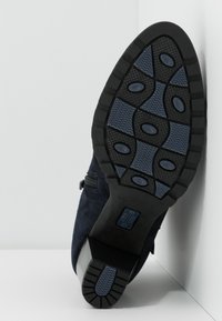 Semelle d’une chaussure noire et bleue avec des motifs de bande de roulement texturés et un talon bloc, positionnée verticalement contre un mur blanc d’angle.