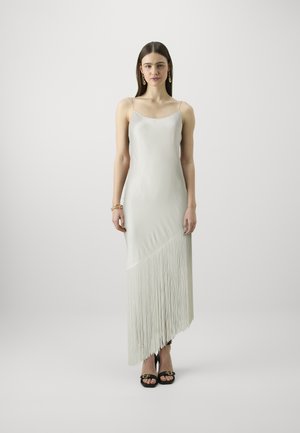 Robe en satin blanc avec des bretelles spaghetti réglables, ourlet asymétrique avec des détails en frange, et une silhouette épurée. Sandales à talons noires visibles.