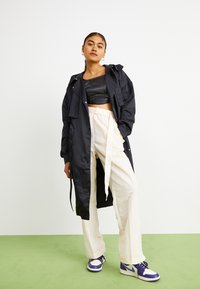 Manteau long noir à finition satinée, associé à un haut court noir, un pantalon large crème et des baskets avec des accents violets.