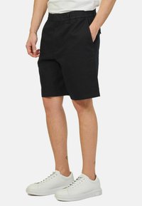 Pantaloni corti neri in cotone con elastico in vita e tasche laterali, abbinati a sneakers bianche. I pantaloni corti hanno una silhouette dritta.