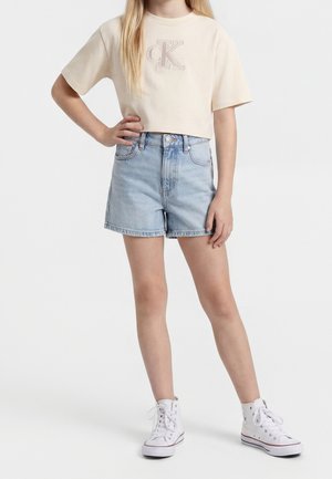 Ung person iført en beige cropped CK t-shirt, lyseblå højtaljede denimshorts og hvide høje sneakers, står med den ene hånd på hoften.