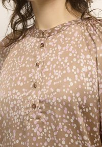 Brown blouse with a textured surface, featuring pink and white polka dots, a rounded neckline, and button detailing down the front.