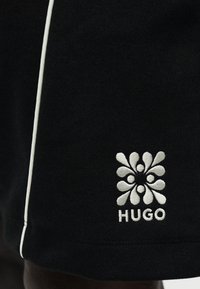 Μαύρο ύφασμα με λευκό κεντητό αφηρημένο λογότυπο ανθών και το κείμενο "HUGO", με μια λεπτή λευκή κάθετη λωρίδα στην αριστερή άκρη.