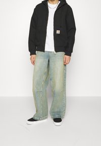 Schwarzer Kapuzenpullover mit Reißverschluss und Fronttaschen, getragen über einem weißen T-Shirt. Hellblaue Jeans mit weitem Bein, kombiniert mit schwarzen und weißen Turnschuhen.