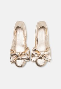 Fémes arany slip-on cipők, melyek elöl egy nagy masnival rendelkeznek, sima felülettel, lekerekített orral, és oldalt ráncolt anyaggal.