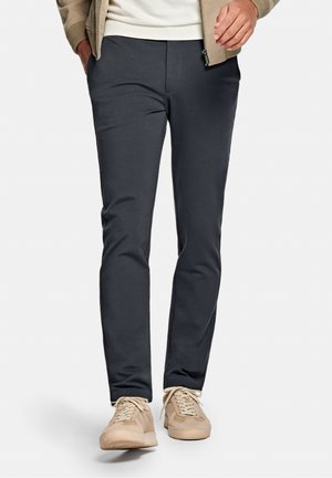 Homme portant un pantalon slim gris foncé, une veste beige et des baskets beiges, marchant les mains partiellement dans les poches sur un fond blanc.