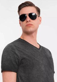 Černé pilotní sluneční brýle, kulaté čočky, tenký kovový rám. Model má na sobě přiléhavé černé tričko s výstřihem do V a malou kapsou na levé straně.