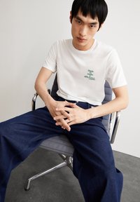 Weißes Baumwoll-T-Shirt mit grünem Text, auf einem silbernen Stuhl sitzend, kombiniert mit dunkelblauen, weit geschnittenen Hosen. Einfaches, lässiges Design, minimaler Stil.