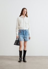 Γυναίκα ντυμένη με λευκό μπουφάν με φερμουάρ, σορτς από τζιν, μαύρες μπότες μέχρι τη μέση της γάμπας, κρατώντας μικρή μαύρη τσάντα, στέκεται σε γκρι δάπεδο μπροστά σε λευκό τοίχο.