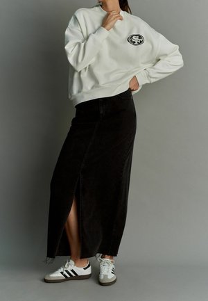 Sweatshirt blanc avec logo, jupe maxi en denim noir avec une fente sur le côté, et baskets blanches avec rayures noires et accents gris.
