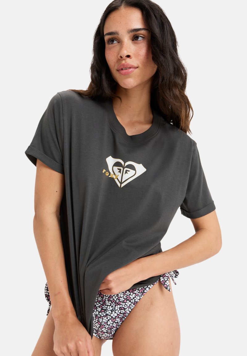 Grå kortærmet t-shirt med et hvidt og gyldent hjerte-logo. Båret over blomsteret mønstrede badedragtsbukser. Blødt, let stof.