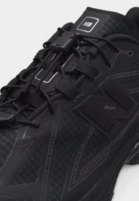 Κοντινό πλάνο μαύρου αθλητικού παπουτσιού με εξαγωνικό δίχτυ, ιμάντες βέλκρο, ελαστικά κορδόνια και λογότυπο "NB" στη μαξιλαρωμένη γλώσσα.