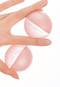 Deux boîtes rondes roses de masque à lèvres au collagène PDRN d’Abib, tenues entre les doigts, présentant une surface lisse et brillante ainsi qu’un design minimaliste.