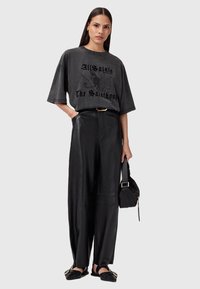 T-shirt grafico oversize nera con stampa "All Saints The Sainthood", abbinata a pantaloni in pelle nera a vita alta e scarpe con tacco nere.